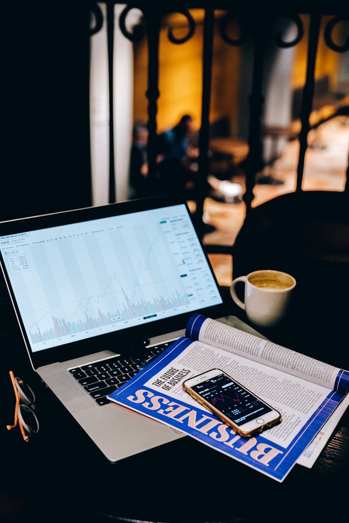 A laptop showing financial charts beside a business magazine and coffee cup in a cafe setting.